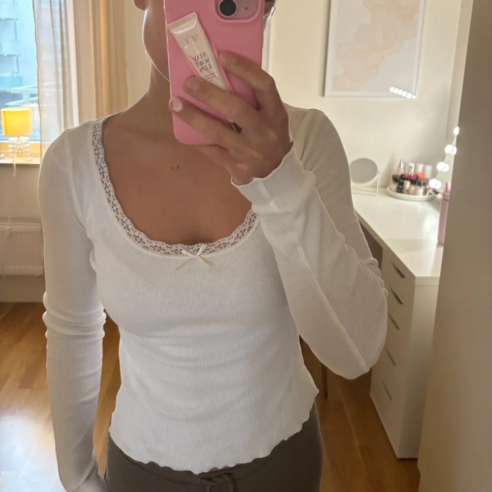 Säljer en stilren vit ribbad topp med långa ärmar och spetsdetaljer vid halsringningen. Toppen har en liten rosett framtill som ger en söt touch. Perfekt för en enkel och elegant look.. Puserot.