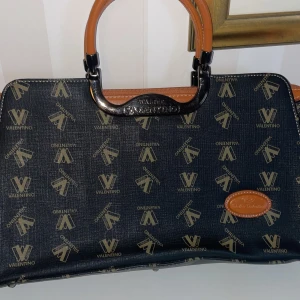 Mönstrad handväska/axelrem från Walter Valentino - Snygg handväska/axelrem  från Walter Valentino med ett unikt mönster i svart och beige. Väskan har bruna handtag och detaljer i skinn. Den har en praktisk dragkedja upptill och är perfekt för att bära dina dagliga nödvändigheter.