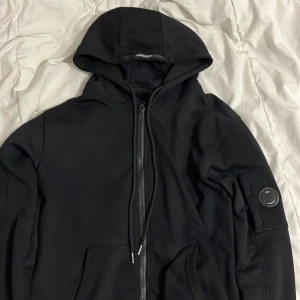 Svart hoodie från C.P. Company - Snygg svart hoodie från C.P. Company med dragkedja och huva. Den har en cool detalj med en rund logotyp på ärmen. Perfekt för en stilren och avslappnad look.