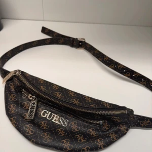  midjeväska från Guess - Snygg svart midjeväska från Guess med ett mönster i brunt och guld. Väskan har en dragkedja och justerbart bälte. Perfekt för att hålla dina saker säkra och stiliga på språng.