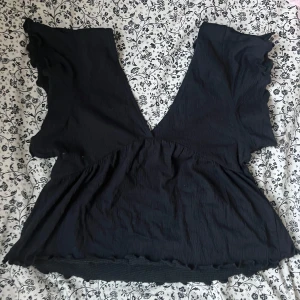 Svart v-ringad topp - Säljer en svart topp med v-ringning och volangdetaljer. Toppen har en lös passform och är perfekt för en stilren look. Materialet är mjukt och bekvämt.