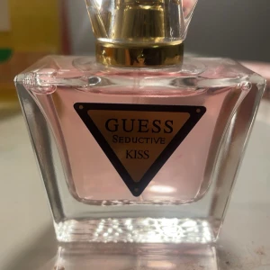 Guess Seductive Kiss Parfym - Säljer en elegant parfym från Guess, Seductive Kiss. Flaskan är i glas med en ljusrosa vätska och har en stilren design med en gyllene kork. Perfekt för den som vill ha en doft som utstrålar både elegans och charm.