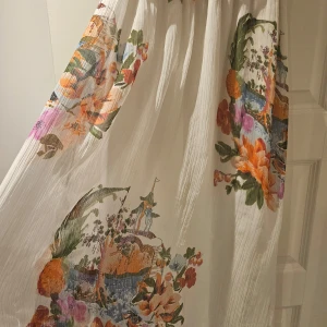 Vit långklänning med färgglatt mönster från Pure - Säljer en vacker vit långklänning från Pure med ett färgglatt, blommigt mönster. Klänningen har en v-ringad design och flätade axelband som ger en bohemisk känsla. Perfekt för somriga dagar och speciella tillfällen.