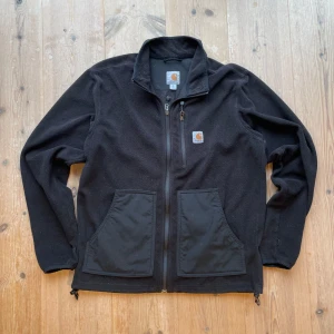 Svart fleecejacka från Carhartt - Säljer en svart fleecejacka från Carhartt med dragkedja och två stora fickor framtill. Jackan har en hög krage och är perfekt för kyligare dagar. Den har en enkel och stilren design med Carhartt-loggan på bröstet samt justerbar midja. Riktigt vintage, skriv vid frågor!