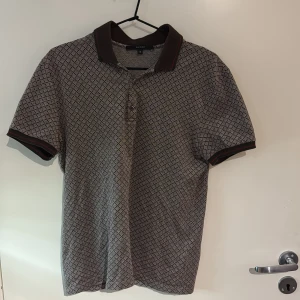 Mönstrad pikétröja från Gucci - Säljer en stilren pikétröja från Gucci med ett diskret mönster i beige och brunt. Tröjan har korta ärmar och en klassisk krage med knappar framtill. Perfekt för en avslappnad men ändå elegant look. (M) sitter som en s för att den är slimfit 