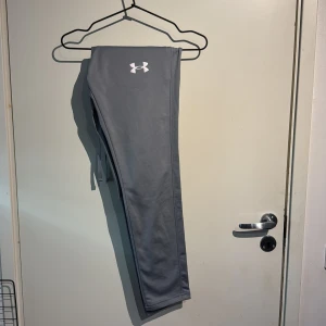 Grå träningsbyxor från Under Armour - Snygga grå träningsbyxor från Under Armour med elastisk midja och dragsko. Byxorna har en diskret logga på framsidan och är perfekta för träning. De är tillverkade i ett bekvämt syntetmaterial som ger bra rörelsefrihet.