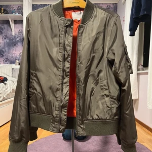 Olivgrön bomberjacka med orange foder - Säljer en olivgrön bomberjacka från Kappahl med ett coolt orange foder. Jackan har dragkedja framtill och en ficka med dragkedja på ärmen.  Jackan är i storlek 160 men jag skulle säga att den är som en S. 