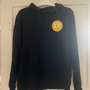 Svart hoodie från Clique - Säljer en svart hoodie från Clique med Hagströmska Gymnasiet-tryck i gult. Tröjan har en klassisk design med huva och dragsko. Perfekt för en avslappnad stil.