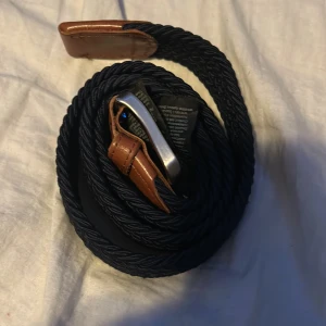 Flätat bälte med läderdetaljer - Snyggt flätat bälte i mörkblått med bruna läderdetaljer och ett stilrent silverfärgat spänne. Perfekt för att ge en extra touch till din outfit,den passar inte mig så jag vill bli av med den med.