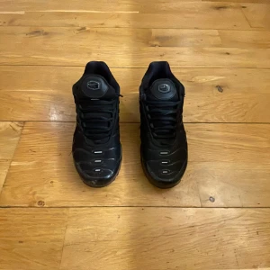 Svarta  Nike tns - Säljer ett par svarta Nike sneakers med en robust design och synlig luftdämpning i sulan. Skorna har snörning och en sportig look, perfekt för en casual stil.