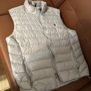 Grå dunväst från Polo Ralph Lauren - Säljer en stilren grå dunväst från Polo Ralph Lauren. Västen har en dragkedja framtill och två praktiska fickor med dragkedjor. Perfekt för lager-på-lager under kyliga dagar.