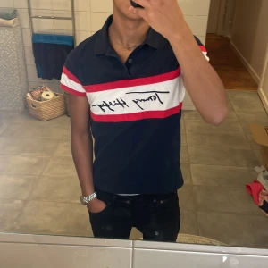 Mörkblå pikétröja från Tommy Hilfiger - Snygg mörkblå pikétröja från Tommy Hilfiger med korta ärmar och röd-vit randig detalj över bröstet. Tröjan har en klassisk krage och knappar framtill. Perfekt för en stilren look! Pris kan diskuteras 