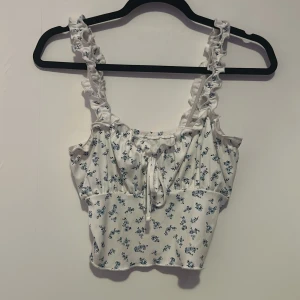 Vit blommig linnetopp med volangdetaljer -  Vit linnetopp med blå blommönster och volangdetaljer på axelbanden. Toppen har en söt knytning framtill och är croppad.