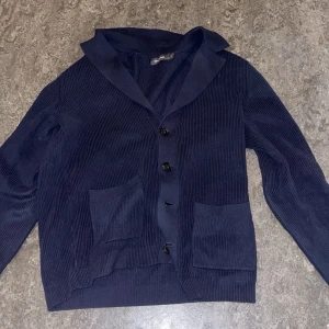 Mörkblå kofta från Dressmann - Säljer en snygg mörkblå kofta från Dressmann Koftan har en ribbad struktur och knappar framtill. Den har två praktiska fickor och en klassisk krage. Perfekt för kyligare dagar. Jätte bra skick