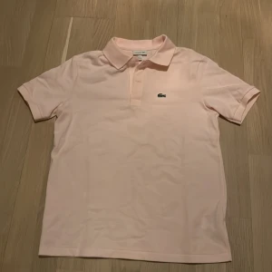 Ljusrosa pikétröja från Lacoste - Säljer en stilren ljusrosa pikétröja från Lacoste. Tröjan har en klassisk krage och korta ärmar med det ikoniska krokodilmärket broderat på bröstet. Perfekt för en avslappnad och snygg look. Storlek 158-164. Ny pris 899 mitt pris 499