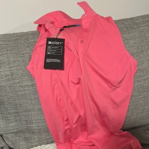 Rosa ärmlös polotopp från J. Lindeberg - Säljer en snygg rosa ärmlös polotopp från J. Lindeberg. Perfekt för en sportig look med knappar framtill och en stilren design. Toppen är i en härlig pop pink färg och passar utmärkt för varmare dagar.