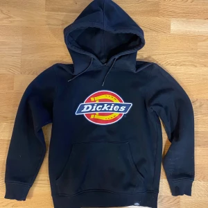 Svart hoodie från Dickies - Säljer en svart hoodie från Dickies med det klassiska logotyptrycket på framsidan. Tröjan har en stor ficka framtill och en bekväm huva. Perfekt för en avslappnad stil.