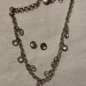 Halsband och örhängen med lila stenar - Elegant halsband med lila stenar som hänger längs kedjan. Matchande örhängen med lila stenar i en rund infattning. Perfekt för att ge en subtil färgklick till din outfit. 
