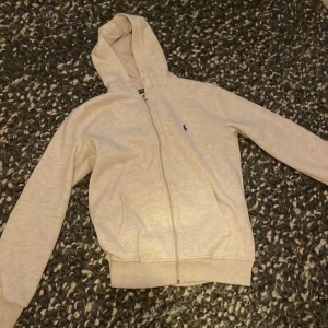 Ralph lauren zipp hoodie - Säljer en stilren beige huvtröja från Polo Ralph Lauren. Tröjan har en dragkedja framtill och en klassisk logga på bröstet. Perfekt för en avslappnad look.