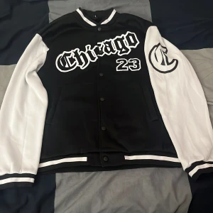 Svart och vit bomberjacka med Chicago-tryck - Snygg svart och vit bomberjacka med tryck av 'Chicago' och siffran '23' på framsidan. Jackan har vita ärmar och svarta knappar framtill. Perfekt för en sportig look.