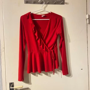 Röd omlottopp från Gina Tricot - Snygg röd omlottopp från Gina Tricot med volangdetaljer och knytband i midjan. Toppen har långa ärmar och en v-ringad design som ger en elegant look. Perfekt för att piffa upp garderoben!