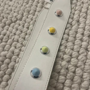 Vitt bälte med färgglada nitar från Gina Tricot - Snyggt vitt bälte från Gina Tricot dekorerat med runda spetsiga nitar i fina pastellfärger . Väsk bandet har en praktisk karbinhake för enkel användning.