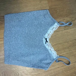 Grå ribbad linnetopp med spetsdetalj - Snygg grå ribbad linnetopp med spetsdetalj vid halsringningen och en liten svart rosett. Perfekt för en stilren och bekväm look.