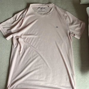 Ljusrosa t-shirt från Calvin Klein - Säljer en stilren ljusrosa t-shirt från Calvin Klein Golf. T-shirten har en liten broderad 'ck' logga på bröstet och är tillverkad i ett mjukt material. Perfekt för en avslappnad look.