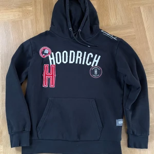 Svart hoodie från Hoodrich - Säljer en svart hoodie från Hoodrich med tryck och broderade detaljer i vitt och rött. Den har en stor ficka framtill och justerbar huva med snören. 
