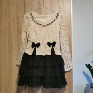 (S) Spetsklänning med pärldetaljer - Jättesöt krämfärgad klänning med svart volangkjol från 5+Angel. Dekorerad med pärlor vid halsringningen samt söta rosetter i midjan. (väldigt kort!) 