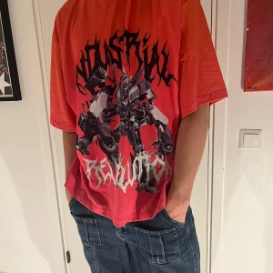 Orange t-shirt med tryck från Jaded London - Cool orange t-shirt från Jaded London med ett stort tryck av robotar och texten 'Revolution'. Perfekt för dig som vill sticka ut med en unik stil. T-shirten har en normal passform och långa ärmar.