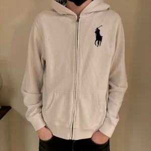 Polo Ralph Lauren zip - Säljer en stilren vit hoodie från Ralph Lauren med dragkedja och det klassiska logotypbroderiet på bröstet. Tröjan har långa ärmar och en bekväm passform, perfekt för en avslappnad stil. Huvan är fodrad med ett randigt mönster i blått och vitt. Nypris är 1999. Hör av dig vid några frågor. 