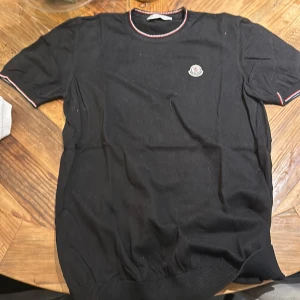 Svart kortärmad tröja från Moncler - Snygg svart tröja från Moncler med korta ärmar och ribbade kanter. Den har en stilren design med röd och vit randdetalj vid hals och ärmslut samt Monclers logga på bröstet. skam funkar kan gå ner i pris med snabba affär