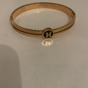 Guldarmband med H-logga - Elegant guldarmband med en stilren design och en framträdande H-logga i mitten. Armbandet har en slät yta med dekorativa detaljer längs kanterna. 