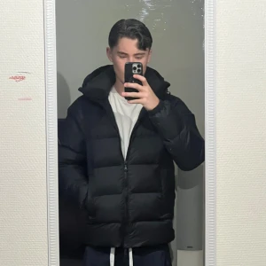 Svart pufferjacka från J.Lindeberg - Säljer en stilren svart pufferjacka från J.Lindeberg. Jackan har en praktisk dragkedja och en mysig huva för extra värme. Perfekt för kyliga dagar.