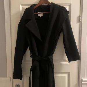 Svart kappa med bälte från VILA - Elegant svart kappa från VILA med knytbälte i midjan. Den har långa ärmar och en klassisk krage, perfekt för höst och vår. Stilren design som passar till många tillfällen.
