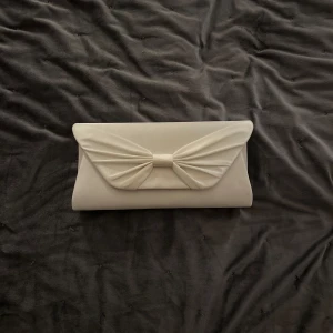 Vit kuvertväska med rosett - Elegant vit kuvertväska med en stilren rosettdetalj på framsidan. Perfekt för speciella tillfällen när du vill ha en chic och minimalistisk look.