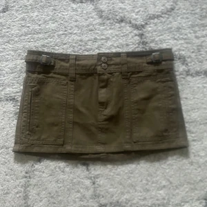 Olivgrön kortkjol med fickor - Snygg olivgrön kortkjol med praktiska fickor och bälteshällor. Kjolen har en knapp- och dragkedjestängning framtill och är perfekt för en avslappnad stil. Den är tillverkad i ett slitstarkt material som ger en cool look.