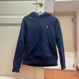 Mörkblå hoodie från Ralph Lauren - Säljer en stilren mörkblå hoodie från Ralph Lauren med det klassiska logotypbroderiet i gult på bröstet. Tröjan har en bekväm passform med långa ärmar och en praktisk magficka. Perfekt för en avslappnad look.