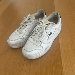 Vita sneakers från Fila - Säljer ett par vita Fila sneakers med snörning. Skorna har en klassisk design med Fila-logga på sidan och hälen. Tillverkade i en kombination av läder och syntetmaterial med textilfoder och EVA/gummisula.