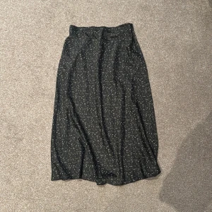 Svart mönstrad kjol från Lindex - Snygg svart långkjol från Lindex med ett diskret mönster i vitt och grönt. Kjolen har en bekväm passform och är perfekt för både vardag och speciella tillfällen. Tillverkad i ett lätt och luftigt material.