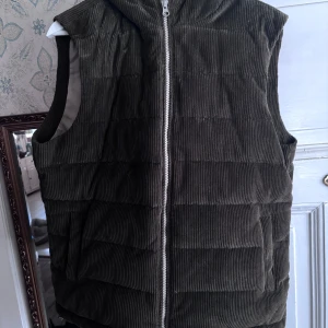 Olivgrön manchester dunväst från Stenströms - Snygg olivgrön dunväst i manchester från Stenströms. Västen har en dragkedja framtill och en mysig huva. Perfekt för kyliga dagar när du vill hålla stilen.