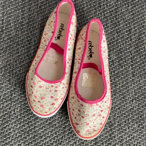 Blommiga ballerinaskor från Rebel - Söta ballerinaskor från Rebel med blommigt mönster i rosa och beige. Skorna har en elastisk rem över foten för bättre passform och en vit sula. Perfekta för en somrig look!
