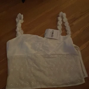 Vit broderad topp från Princess Polly - Säljer en söt vit topp från Princess Polly med broderade detaljer och unika axelband. Perfekt för sommardagar och festliga tillfällen. Toppen har en croppad passform och är tillverkad i ett lätt och luftigt material.