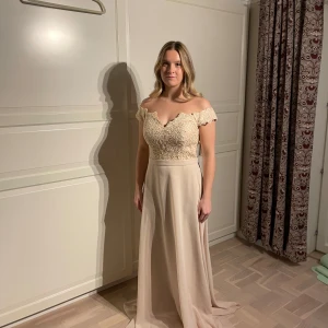 Champagnefärgad långklänning med spetsdetaljer - Elegant champagnefärgad långklänning med offshoulder-design och vacker spets upptill. Klänningen har en snörning i ryggen och ett flytande kjolparti. Perfekt för formella tillfällen.