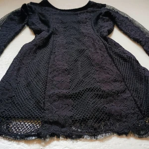 Svart spetsklänning - Elegant svart klänning i spets med långärmad design. Klänningen har ett vackert blommönster och ett nätliknande material som ger en sofistikerad look. Perfekt för speciella tillfällen.