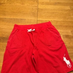 Röda shorts från Polo Ralph Lauren - Snygga röda shorts från Polo Ralph Lauren med en stor vit logga på benet. De har snörning i midjan och en bakficka. Perfekta för en avslappnad stil.