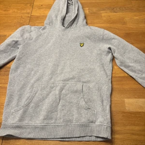 Grå hoodie från Lyle & Scott - Säljer en stilren grå hoodie från Lyle & Scott med deras ikoniska logga på bröstet. Tröjan har en praktisk magficka och en bekväm huva. Perfekt för en avslappnad stil.