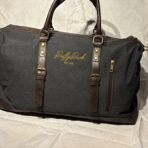 Svart och brun Duffelväska från Duffy Duck - Snygg och rymlig svart duffelväska med bruna läderdetaljer från Duffy Duck. Väskan har två handtag och en dragkedja upptill. Perfekt för resor eller gymmet. Bra fack inuti 