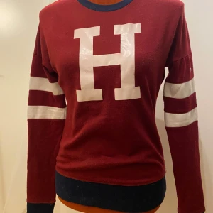Vinröd tröja med bokstaven H från Horseware - Snygg vinröd tröja från Horseware med ett stort vitt H på framsidan. Tröjan har långa ärmar med vita ränder och en mörkblå krage. Perfekt för en sportig look! Långärmad tröja med tryck  Längd: 63 cm Armhåla till armhåla: 48 cm  Inga bilder när den sitter på mig! Katter finns i hemmet 